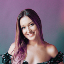A woman with long, straight hair partially dyed purple smiles at the camera, radiating warmth and guidance. She wears an off-the-shoulder, floral-patterned top and poses against a purple gradient background.