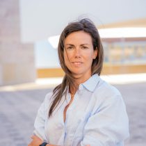 A woman with long brown hair stands outdoors, wearing a light blue button-up shirt. She looks confidently at the camera with a slight smile, embodying the spirit of coaching and guidance. The background is bright and modern, with blurred architectural features.