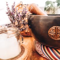 A Sound Healing & Spiritual Guidance set features a small glass candle and dried lavender next to a dark wooden bowl with an engraved symbol, all arranged on a colorful woven mat atop a wooden surface.