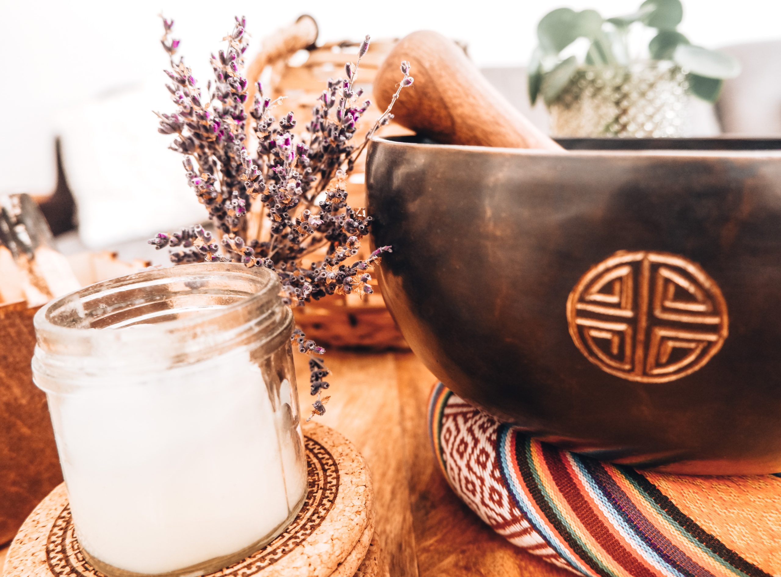 A Sound Healing & Spiritual Guidance set features a small glass candle and dried lavender next to a dark wooden bowl with an engraved symbol, all arranged on a colorful woven mat atop a wooden surface.