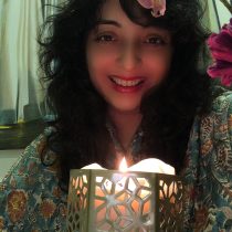 A woman with curly dark hair and a bright smile holds a Tarot Reading & Spiritual Guidance candle. She wears a patterned outfit with an orchid in her hair, and curtains and flowers are in the background.