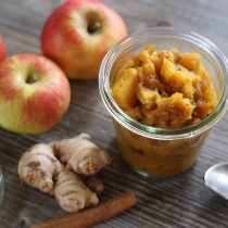 A brochure for Ayurveda Consultation rests on a wooden table alongside fresh apples, ginger root, a cinnamon stick, a spoon, and a jar lid.