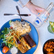 A person enjoys grilled fish, rice, salad, and shrimp next to a cocktail and clear water bottle—a delicious meal highlighting the benefits of the 3-Month Mind + Body Reset personalized nutrition coaching program.