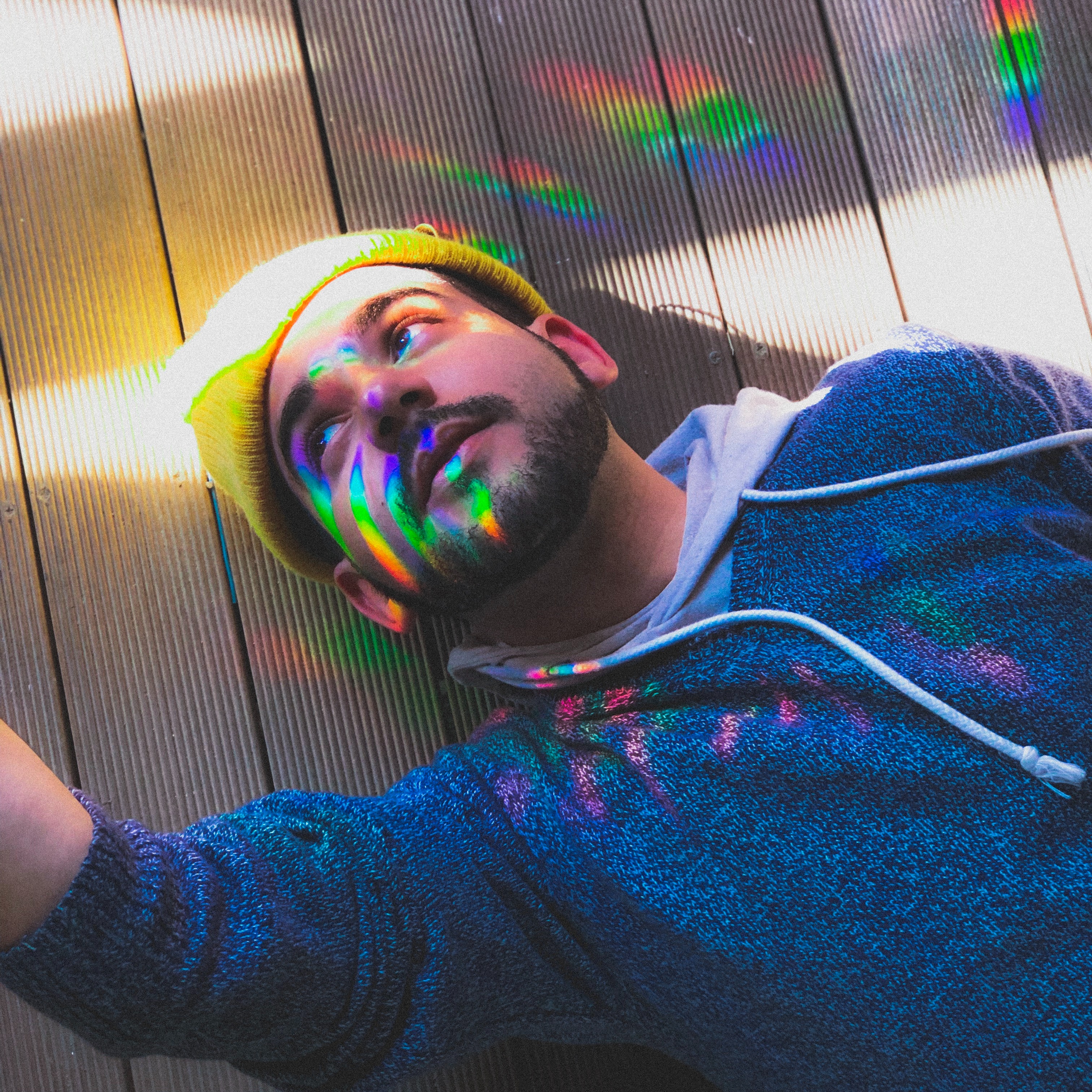 Man wearing a yellow beanie and a layered white hoody under a heather grey sweatsirt is laying on the ground looking up and away. There is rainbow light shining on him as he receives long distance reiki.