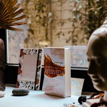 A windowsill displays stones, a brown vase with dried palm leaves, books—including one titled Ayurveda Consultation—and a white Buddha head statue as soft natural light gently filters through the window.