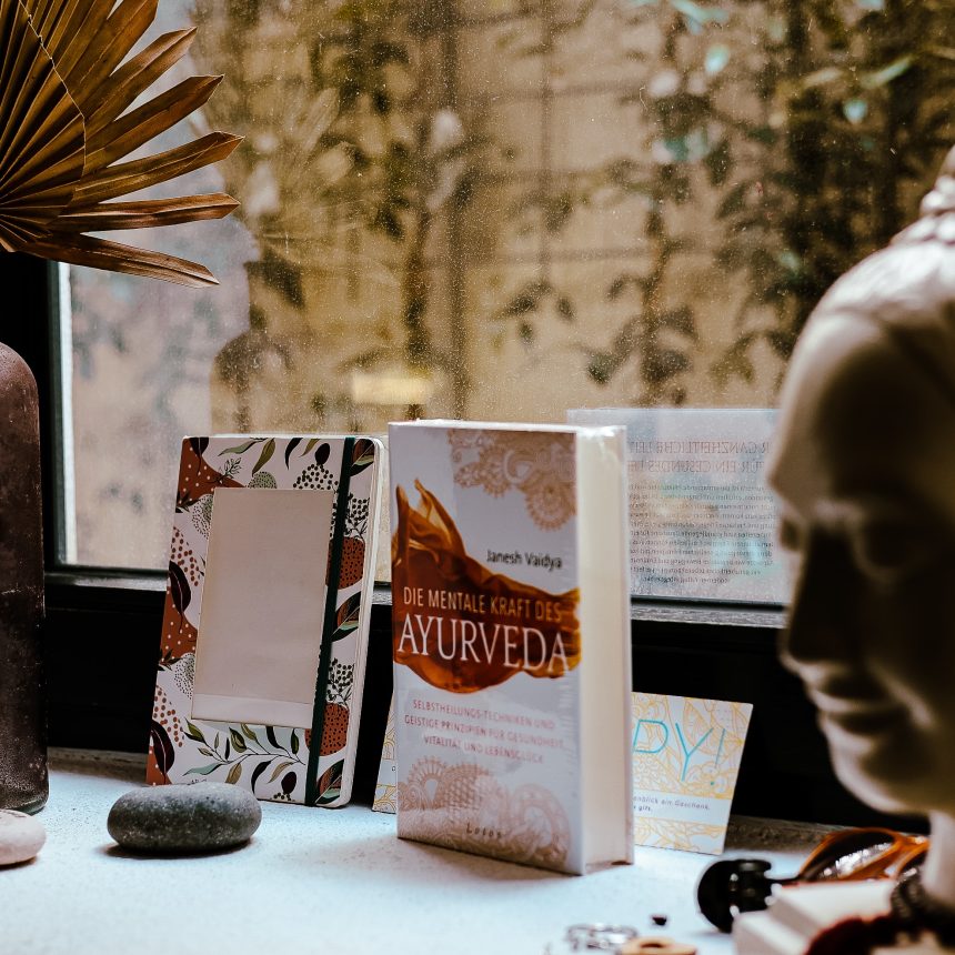 A windowsill displays stones, a brown vase with dried palm leaves, books—including one titled Ayurveda Consultation—and a white Buddha head statue as soft natural light gently filters through the window.