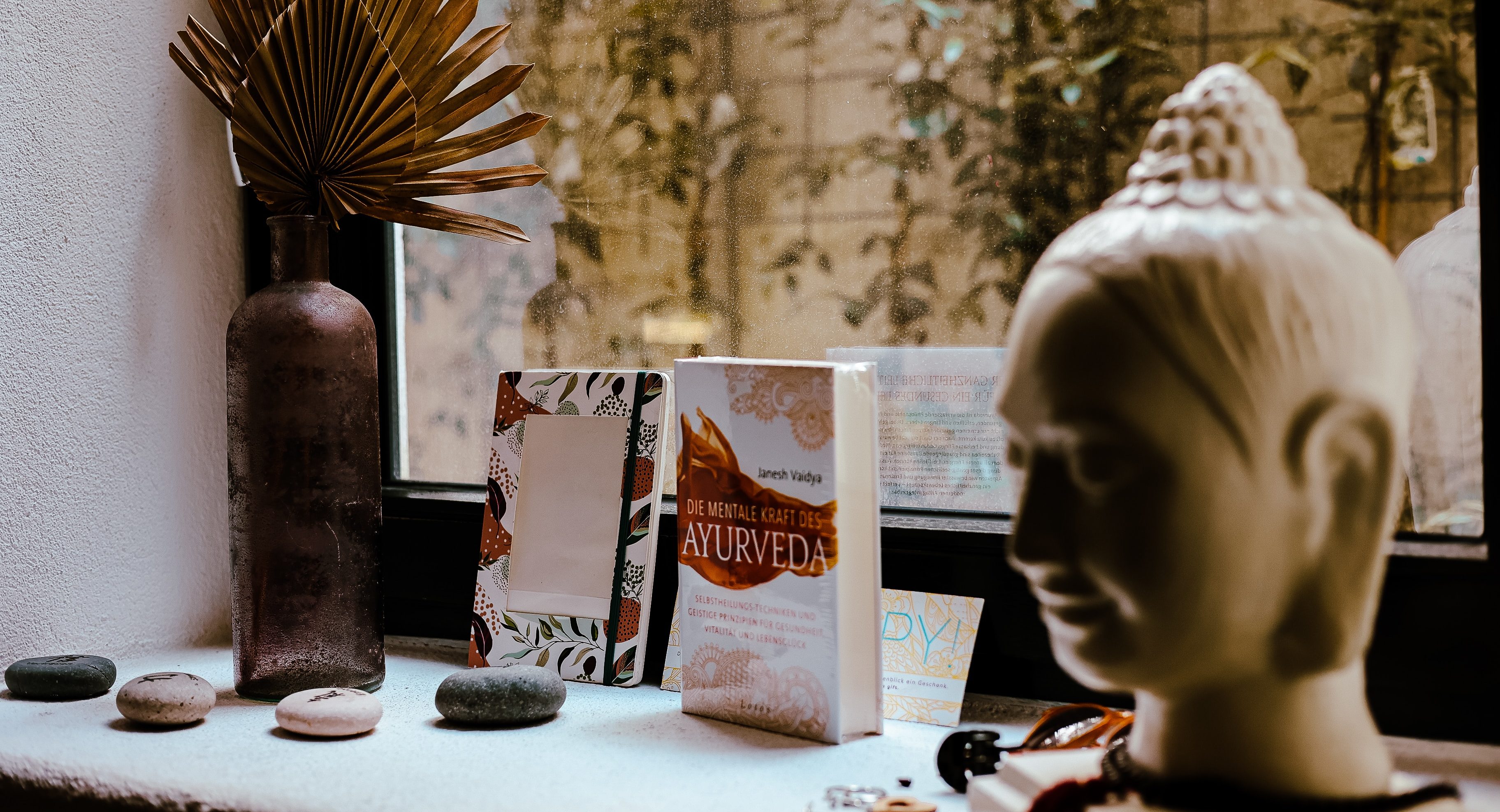 A windowsill displays stones, a brown vase with dried palm leaves, books—including one titled Ayurveda Consultation—and a white Buddha head statue as soft natural light gently filters through the window.