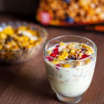 A glass of yogurt, granola, and pomegranate seeds sits on a wooden surface—an inviting scene that hints at the benefits of the 3-Month Mind + Body Reset. In the background, a similar bowl and cereal box are slightly out of focus.