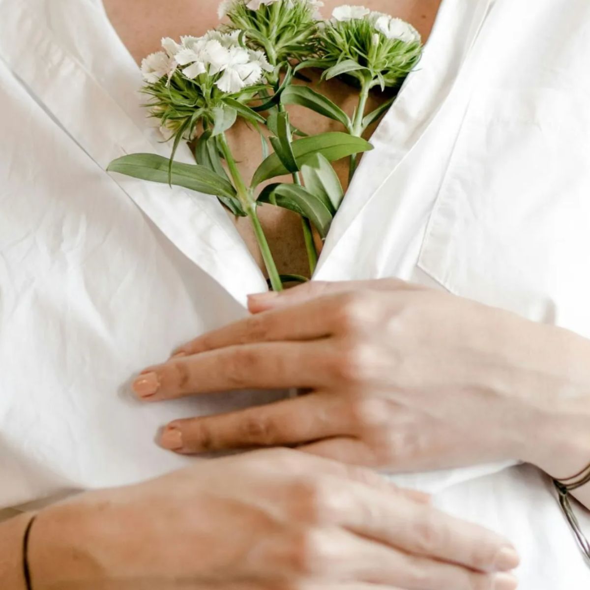 A person in a white shirt, hands over their chest and small white flowers in the open collar, embodies the calm and support offered by the Private Coaching Container.