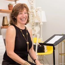During The Shifter Training Session, a woman with brown hair in a sleeveless black dress laughs while seated. Behind her, a skeleton model, yellow chair, music stand with papers, and decorated shelves create an inviting scene.