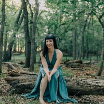 A woman in a flowing teal dress sits barefoot on a forest log, smiling warmly as she invites you to experience the Sound Healing & Intuitive Guidance Package amidst tranquil trees and leaves.