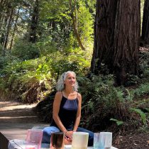 A woman sits cross-legged on a wooden platform in a forest, surrounded by tall trees. She is smiling and has crystal singing bowls placed in front of her on a cloth. Sunlight filters through the leaves above.