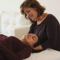 During The Shifter Training Session, a woman gently supports another’s head in a calming, softly lit room with a white couch in the background; both wear dark tops and focus on relaxation and fall prevention.
