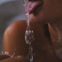 A close-up of a person’s mouth and shoulder as they stick out their tongue to catch a stream of flowing water. The lighting is soft and the focus is on the water and lips.