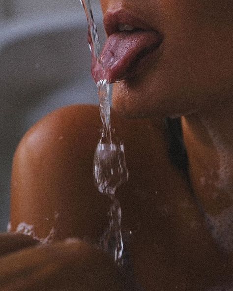 A close-up of a person’s mouth and shoulder as they stick out their tongue to catch a stream of flowing water. The lighting is soft and the focus is on the water and lips.