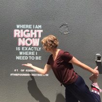 After a Soulful Harmony Flow session, a person strikes a yoga pose, smiling at a wall mural that reads, WHERE I AM RIGHT NOW IS EXACTLY WHERE I NEED TO BE, basking in the sun as their shadow stretches across the wall.