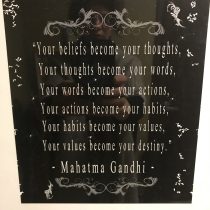 The Soulful Harmony Flow framed plaque features an inspiring Mahatma Gandhi quote on beliefs shaping destiny, with ornate white designs on a black background—an uplifting accent for any yoga space. A faint photographer’s reflection adds subtle character.