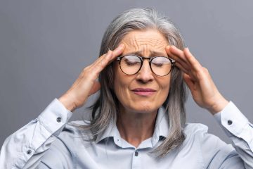 An older woman with gray hair and glasses touches her temples, eyes closed and brows furrowed, appearing to suffer from professional burnout against a plain gray background.