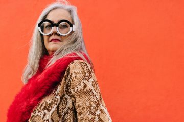 Older woman with long gray hair, wearing large round glasses, a snake-print coat, and a red fuzzy scarf stands confidently in front of a bright orange wall, embodying unconditional acceptance and self-assurance.