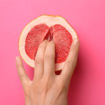 A hand with two fingers parted is pressed into a halved grapefruit on a bright pink background, symbolizing pleasure coaching in the SensualSoul: Empowerment product.