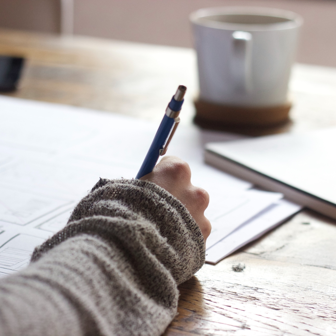 A person in a gray sweater writes at a wooden table with a white mug and closed notebook, organizing support for new mothers using Clarity Plus - Matrescence Coaching + Resources.