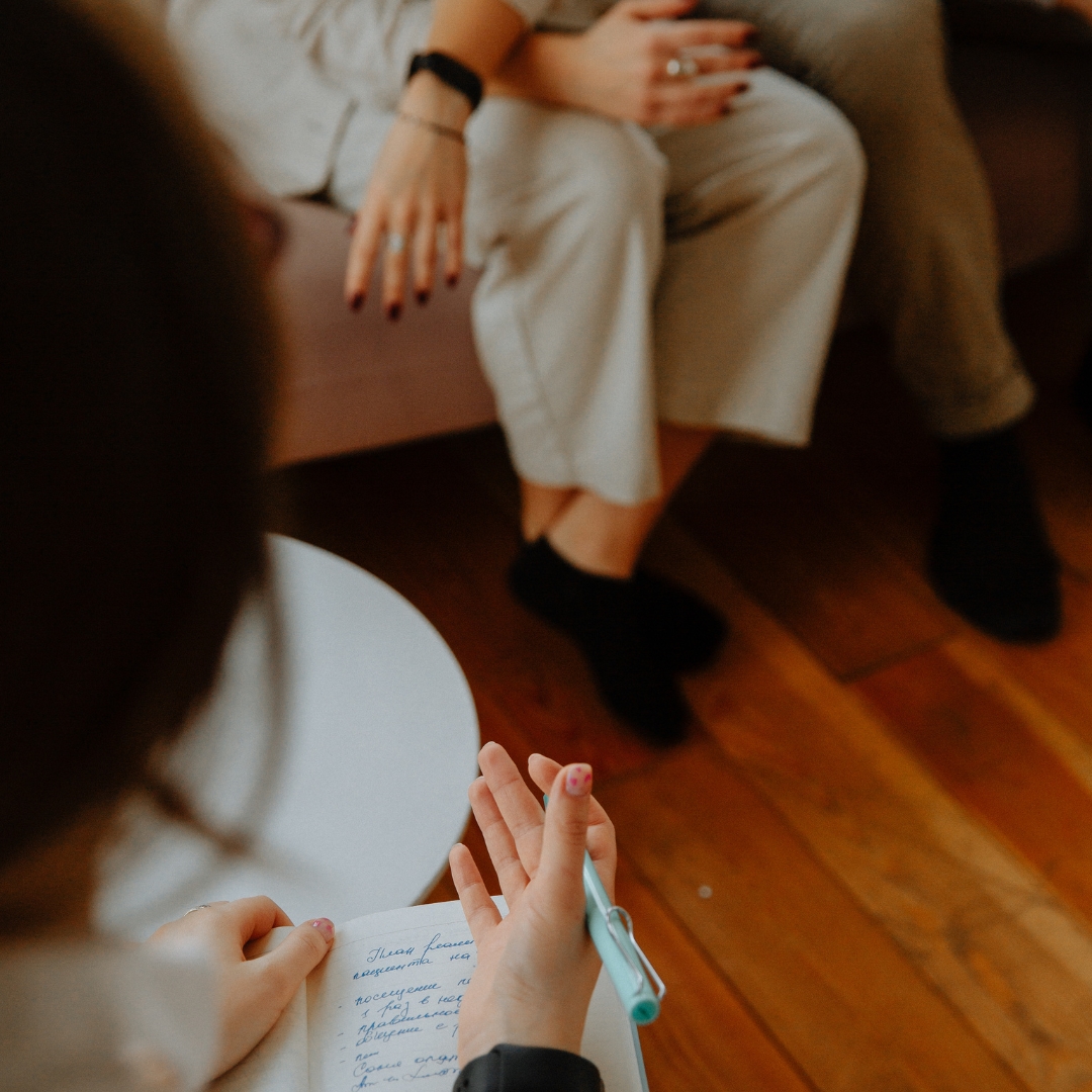 During a Clarity - Matrescence Coaching session, one person sits on a couch with hands folded on their knees while another takes notes in a notebook.