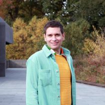 Boshko is wearing a bright yelllow knitted sweater underneath and unbuttoned turquoise corduroy longsleeve shirt. Boshko leads reiki and sound healing sessions for groups.