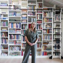 A woman wearing glasses and a green jumpsuit stands in front of a large, tall bookshelf filled with colorful books in a bright room with light wood floors and a high, slanted ceiling.