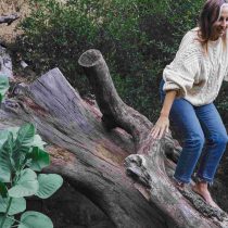 A woman in a cream sweater and blue jeans balances barefoot on a large fallen tree trunk in a green, wooded area.