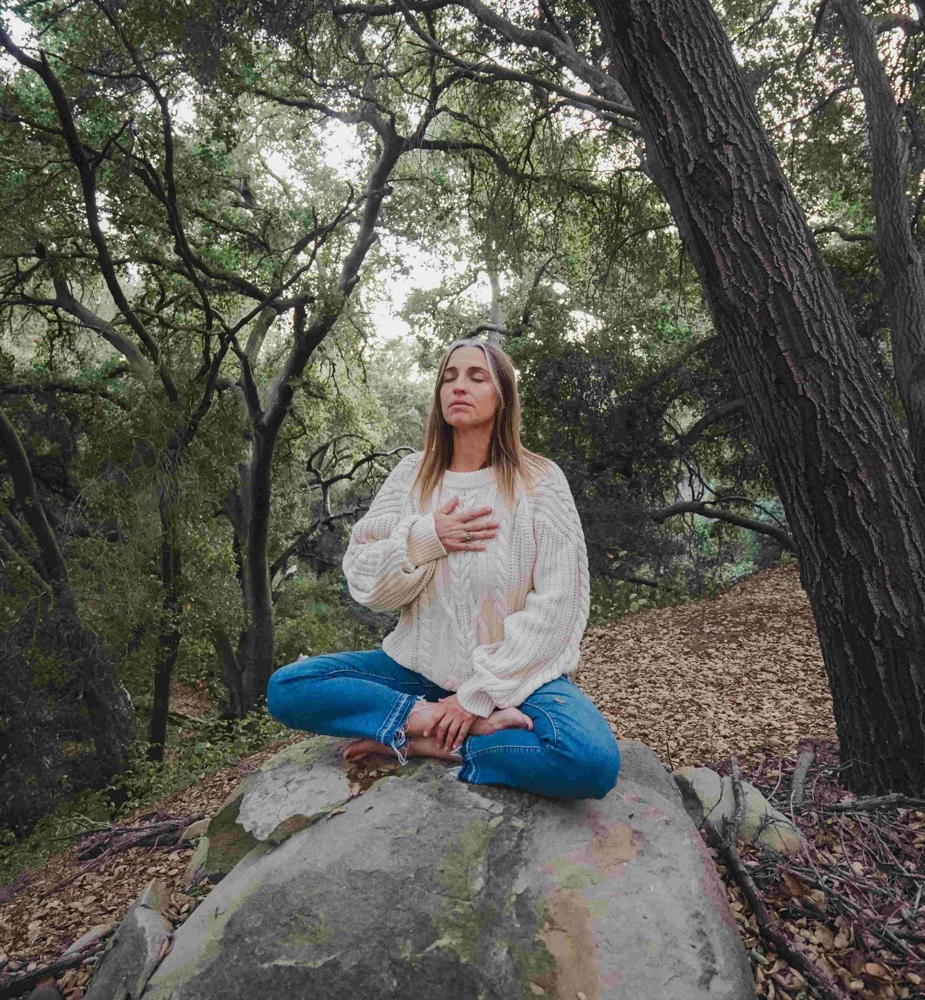 Kate, a coach who helps people find the best way to deal with anxiety, is sitting cross legged on a boulder out in nature. She is wearing a white sweater, blue jeans and no shoes. She has her right hand placed over her heart and her eyes closed.