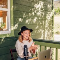 A woman wearing a black hat and light sweater smiles while sitting on a porch, speaking into a microphone with a laptop in front of her on a small wooden table. Sunlight and tree shadows decorate the green wall behind her.