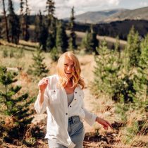 A woman with red hair wearing a white shirt and ripped jeans smiles while walking through a sunlit forested area with mountains and trees in the background.