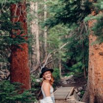 A woman wearing a black hat, light top, and jeans smiles while standing on a forest trail surrounded by tall trees and greenery. A wooden footbridge is visible in the background.
