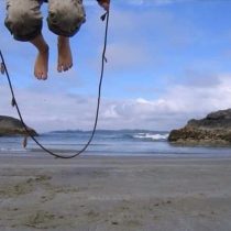 A person jumps barefoot with a skipping rope on a sandy beach, captured mid-air, with the ocean and rocky formations in the background under a partly cloudy sky.
