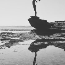 A person stands on a rocky ledge by the sea with arms outstretched, reflected in a calm tidal pool below. The image is in black and white, capturing the silhouette against the sky and water.