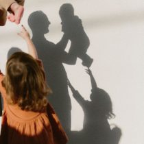 A child in an orange dress reaches toward an adult holding a baby, casting clear shadows of all three figures against a sunlit white wall.
