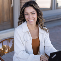 A woman with wavy brown hair, a life purpose coach, smiles at the camera outdoors in a beige jacket and brown top, seated at a table with her laptop while representing the 2-Month Life Purpose Coaching Program.