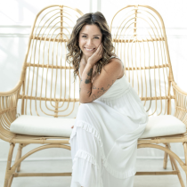 A woman in a white dress sits on a wicker bench, smiling thoughtfully in a bright, naturally lit space—reflecting the calm confidence inspired by the 4-Month Transformational Coaching Program.