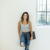 A confident woman with wavy hair in a gray crop top and ripped jeans stands by a barred window, holding a black laptop. Her look embodies the empowerment gained through the 4-Month Transformational Coaching Program.