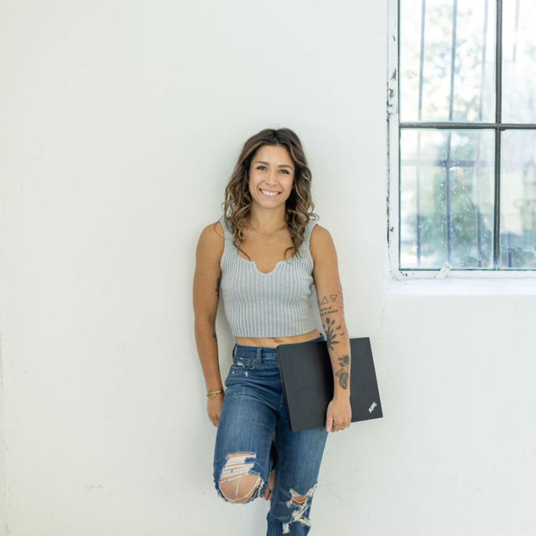 A confident woman with wavy hair in a gray crop top and ripped jeans stands by a barred window, holding a black laptop. Her look embodies the empowerment gained through the 4-Month Transformational Coaching Program.