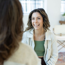 A smiling woman with wavy brown hair, in a light shirt over a green top, discusses the 4-Month Transformational Coaching Program with another person in a bright, relaxed indoor setting.