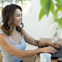A woman with wavy hair and tattoos, a life purpose coach, smiles while typing on a laptop at a wooden table. The space features a framed photo, green plant, white mug, and natural light—showcasing the 2-Month Life Purpose Coaching Program.