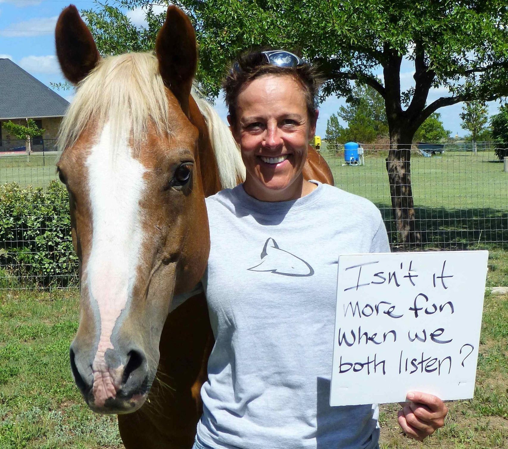 A smiling person stands next to a light brown horse, holding a sign that reads, Isnt it more fun when we both listen? The scene is outdoors with trees, grass, and a fence in the background.
