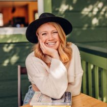 A smiling woman with long blonde hair wearing a black hat and cream sweater sits at an outdoor table, leaning on two books and resting her chin on her hand, with a green wall and sunlight in the background.