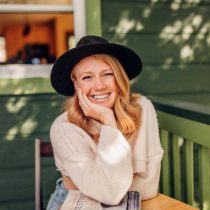 A smiling woman with blonde hair and a black hat sits at a wooden table outside, resting her head on her hand. Three books are stacked in front of her. She is wearing a light sweater and jeans, with a green house in the background.