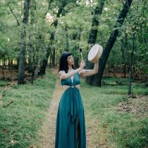 A woman in a long teal dress stands on a forest path holding the Sound Healing & Intuitive Guidance Package, framed by tall green trees and lush grass as if leading a healing session.