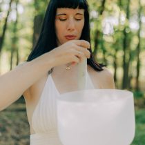 A woman stands among trees, playing a frosted crystal singing bowl with sunlight filtering through the leaves, creating a peaceful scene like those experienced in our Sound Healing & Intuitive Guidance Package.
