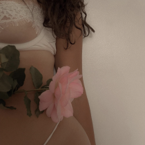 A person wearing white lingerie holds a large pink rose with green leaves against their body, standing in front of a plain light-colored wall. Only the torso and part of the face are visible.