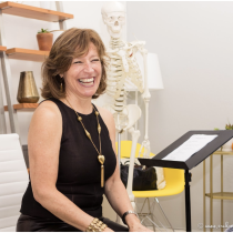 A woman in a black dress smiles in a modern office, with The Burch Method Assessment skeleton model nearby, a music stand with papers, a white chair, and shelves in the background.