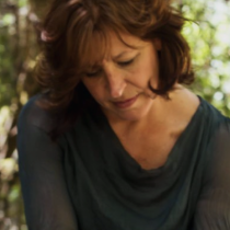 A woman with wavy brown hair in a sheer, dark top looks down thoughtfully in a sunlit, leafy forest—perhaps reflecting after completing The Burch Method Assessment.